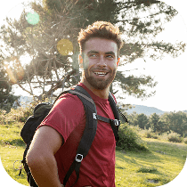 A smiling man wearing a red shirt and a backpack stands outdoors in a grassy area with trees and sunlight in the background.