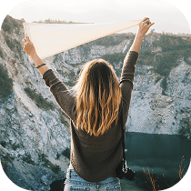 A woman with long, blonde hair stands on a rocky cliff, facing away, holding a white scarf above her head with both hands. She overlooks a scenic landscape with a body of water below.