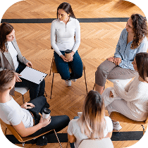 A group of six people sit in a circle on chairs, engaged in conversation, in a bright room with wooden flooring.
