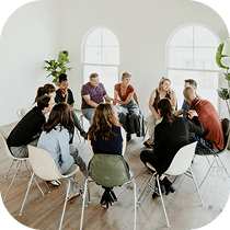A diverse group of people sit in a circle of chairs in a bright room with large windows, engaged in a group discussion or support meeting.