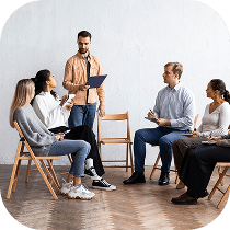 A group of five adults sit in a circle of wooden chairs, attentively listening to a man standing and holding a clipboard, suggesting a discussion or group meeting in a bright room.