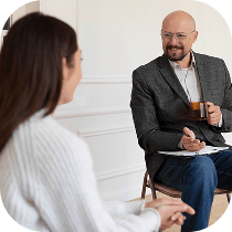 A man holding a cup and clipboard sits across from a woman in a white sweater, engaging in conversation in a bright, minimalist room.