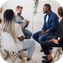 A group of five people sit in a circle indoors, engaged in a discussion. One person holds a clipboard, and everyone appears attentive and involved in the conversation.