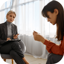 A therapist sits with a notepad, attentively listening to a woman who looks distressed and is seated across from her, holding her hands together.
