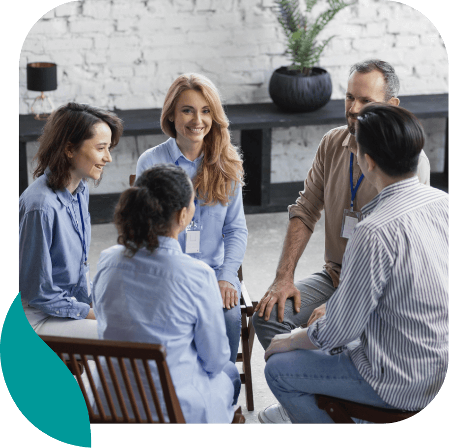 A group of five adults sits in a circle, smiling and talking, in a bright room with a white brick wall and a potted plant in the background. They appear to be participating in a group discussion or support session.