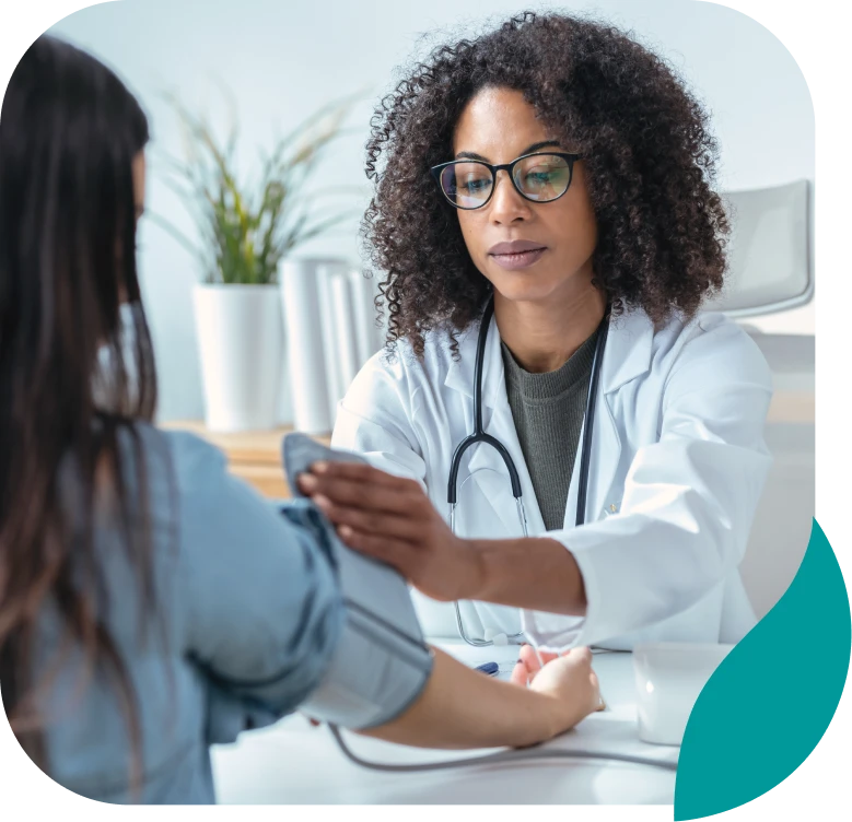 A female doctor in a white coat and glasses checks a patient's blood pressure in a bright medical office with plants in the background.