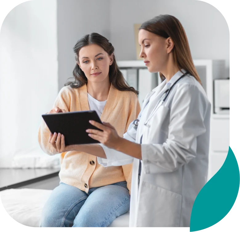 A doctor in a white coat shows a digital tablet to a seated woman in casual clothes, discussing information together in a bright, modern medical office.