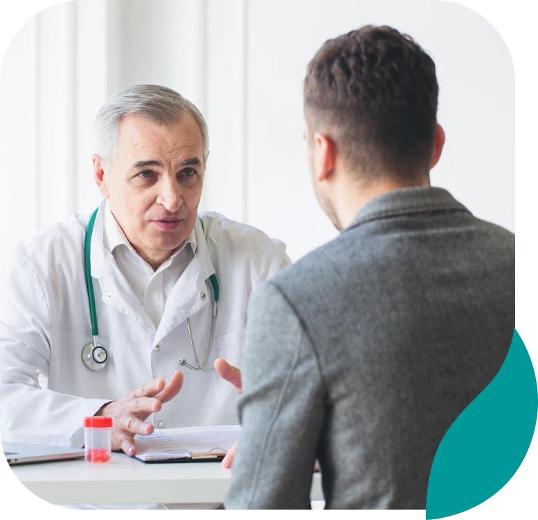 A doctor in a white coat with a stethoscope talks to a patient across a desk. The doctor appears attentive and engaged, while the patient listens. A small medical container sits on the desk between them.