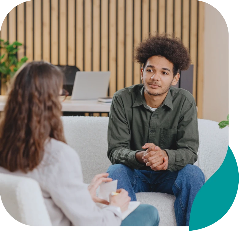 Two people sit across from each other in a modern office, engaged in conversation. One person listens attentively while the other holds a notepad, suggesting a counseling or interview session.